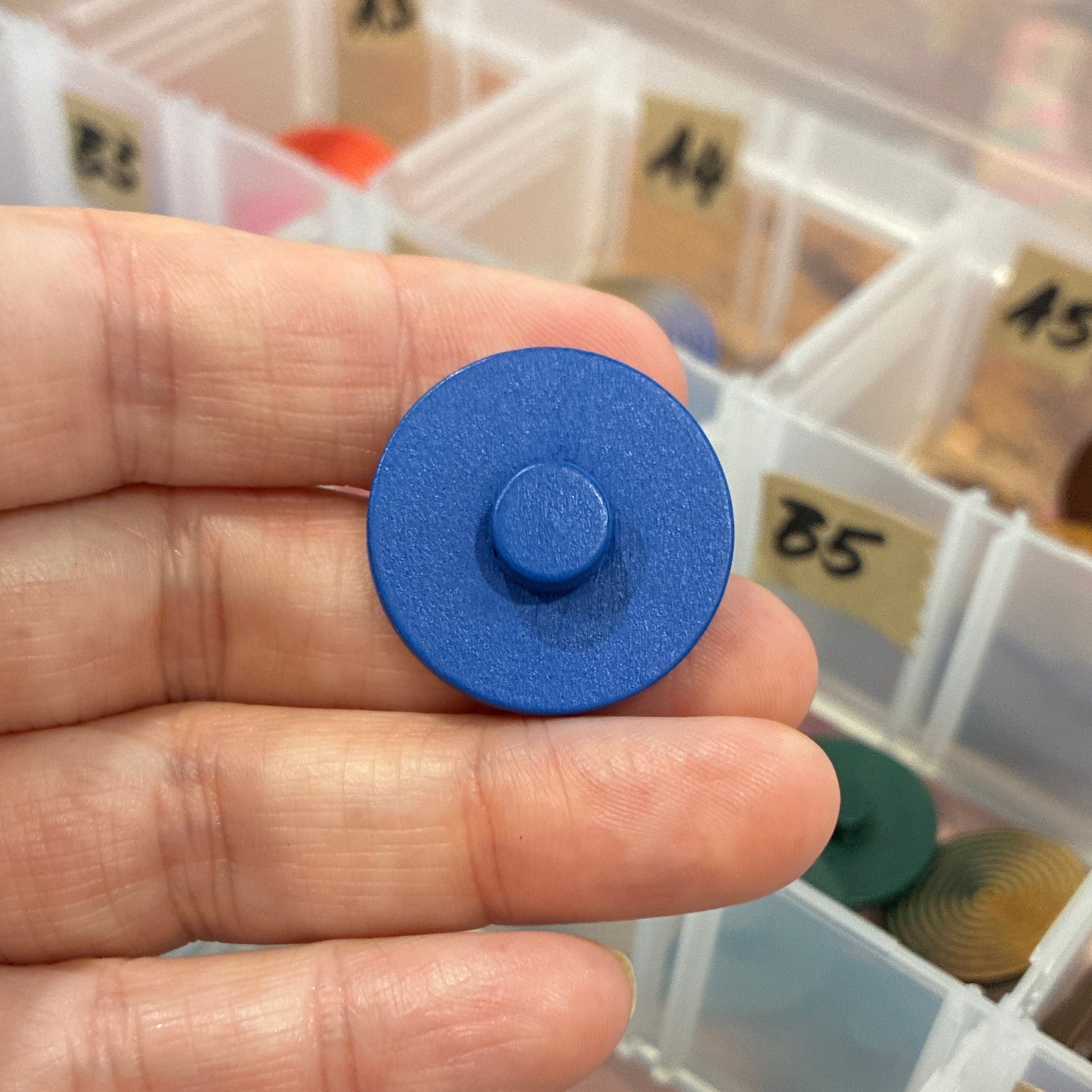 Blue circular button held in a hand with a container of assorted colored objects in the background