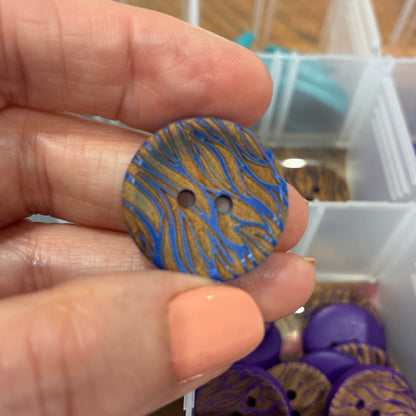 Hand holding a blue and gold patterned button with a container of buttons in the background.