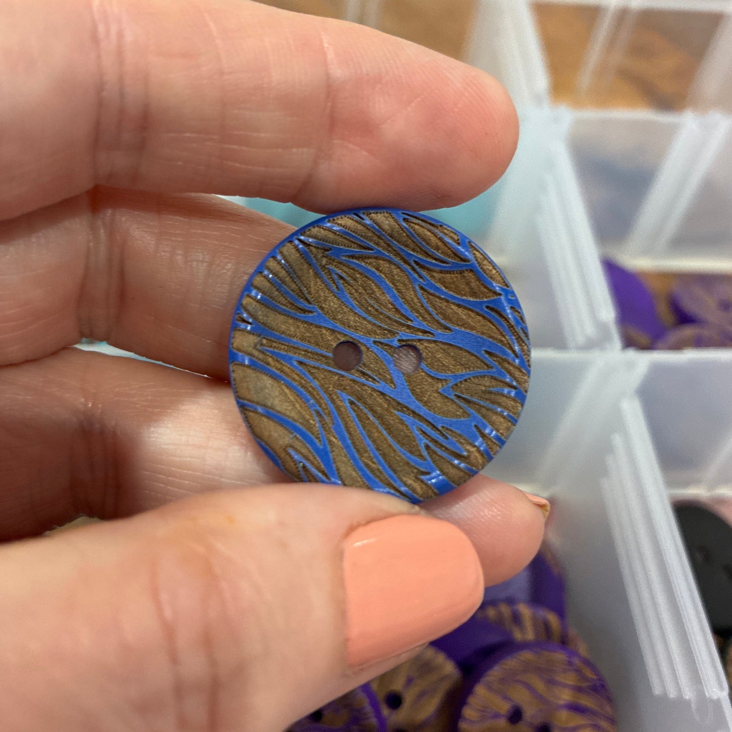 Hand holding a blue patterned button with a blurred background of a storage container.