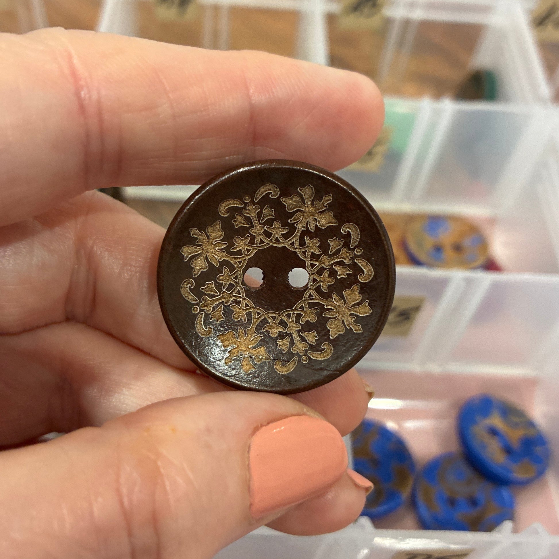 Hand holding a decorative button with intricate designs, blurred background of a container with buttons.