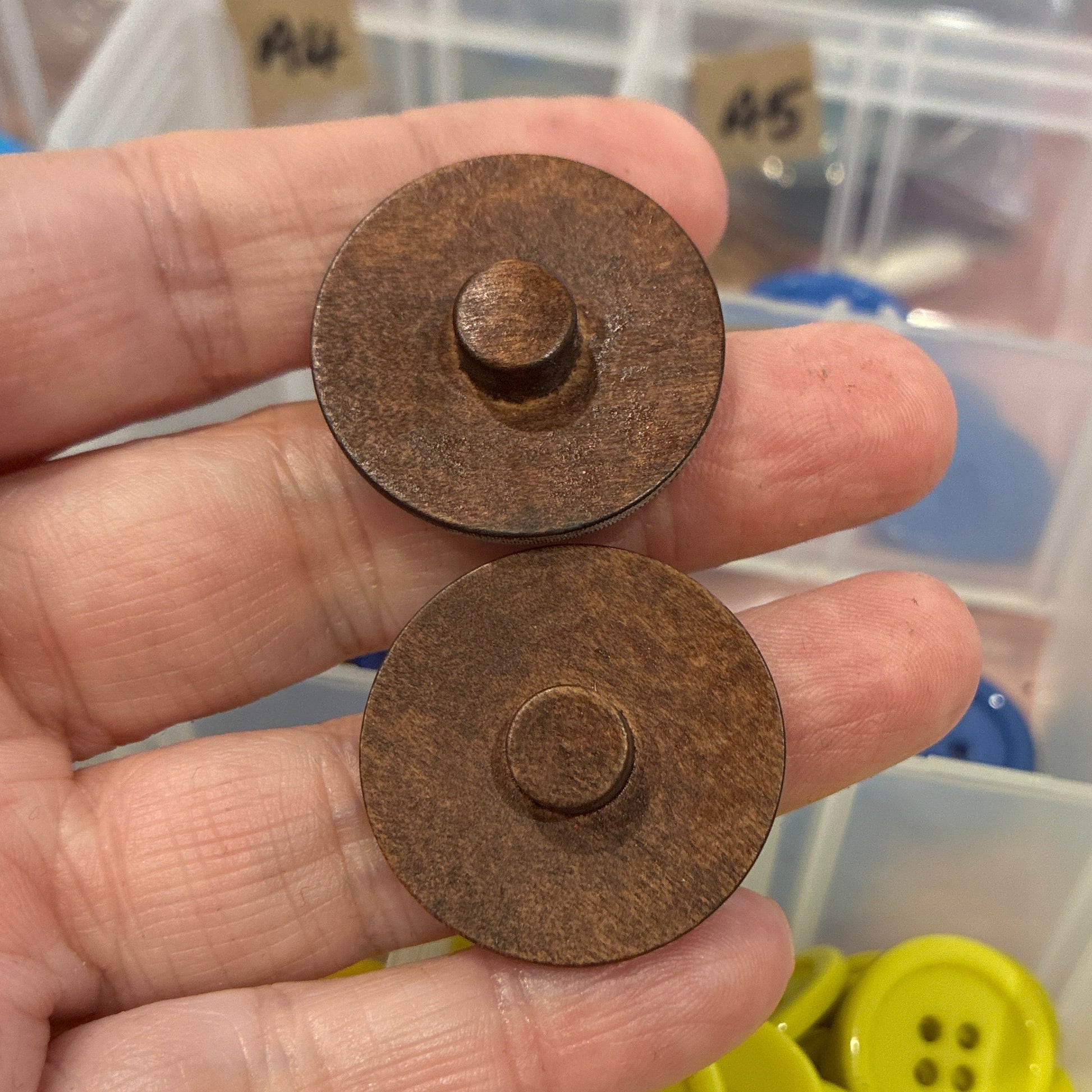 Two wooden buttons held in a hand with a background of colorful buttons in containers.