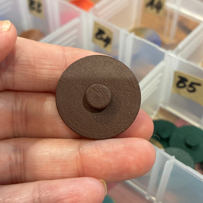 Brown circular button held between fingers with a container of colorful round objects in the background