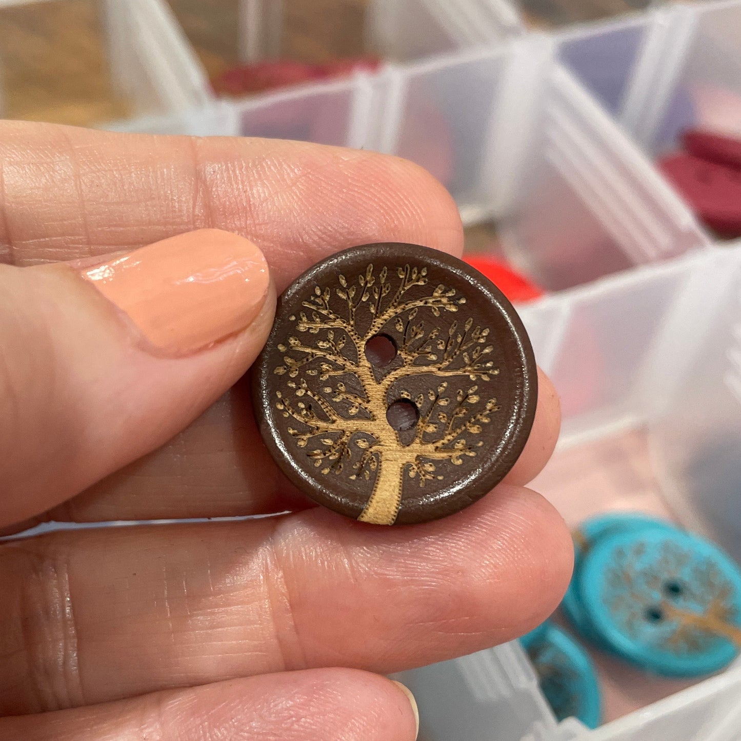 Hand holding a decorative button with intricate design, against a background of a clear container with more buttons.