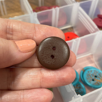 Hand holding a brown button with a clear plastic container of buttons in the background
