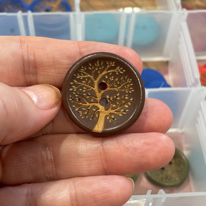 Hand holding a button with a tree design in front of a container of assorted buttons.