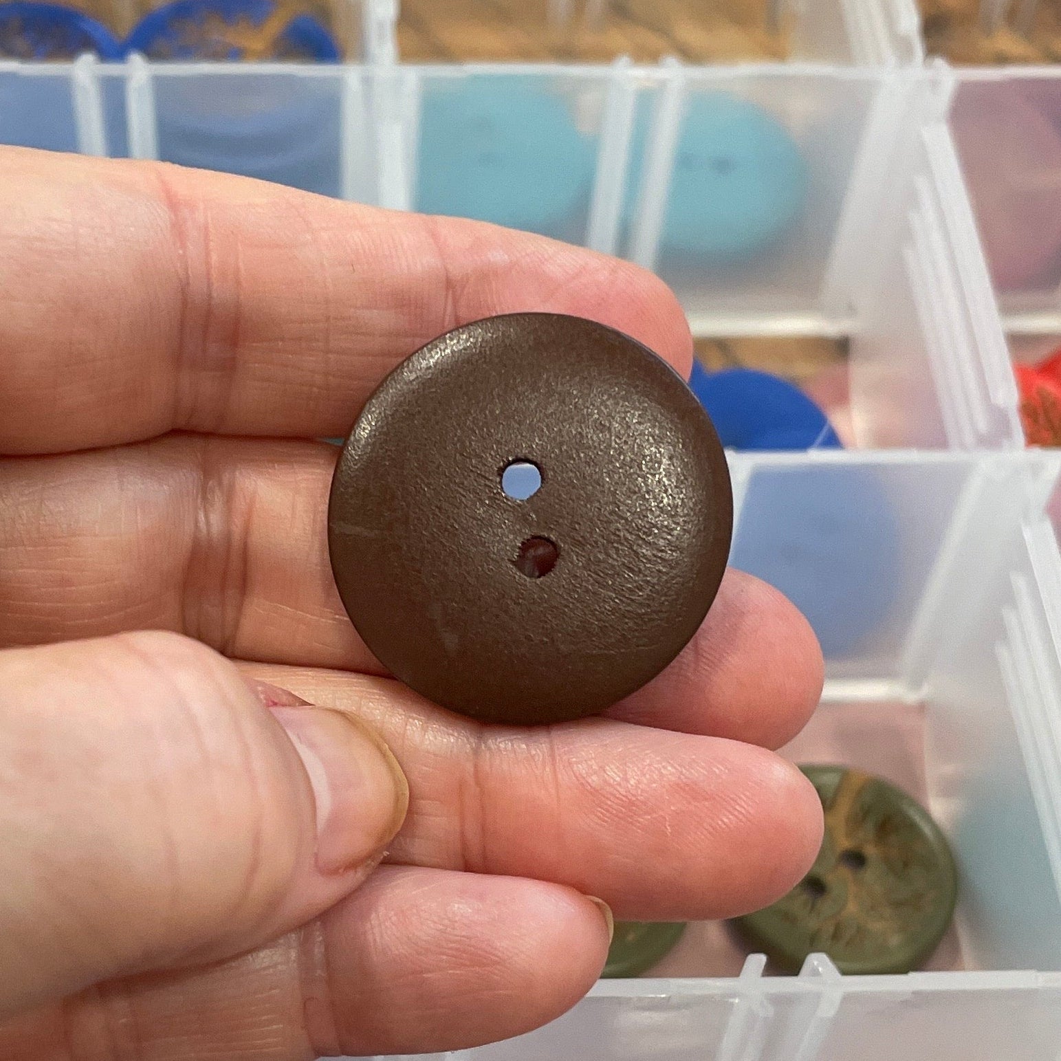 Hand holding a brown button with a container of colorful buttons in the background