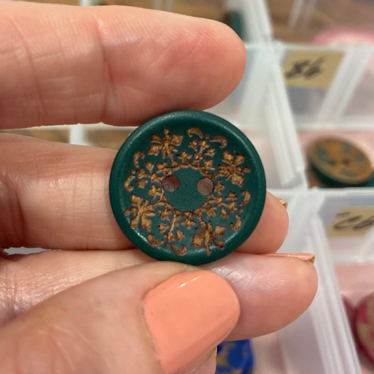 Hand holding a green decorative button with intricate designs, against a blurred background of a storage container.