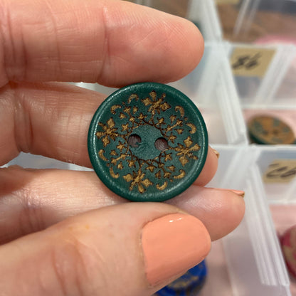Hand holding a decorative button with intricate design in front of a container of buttons.