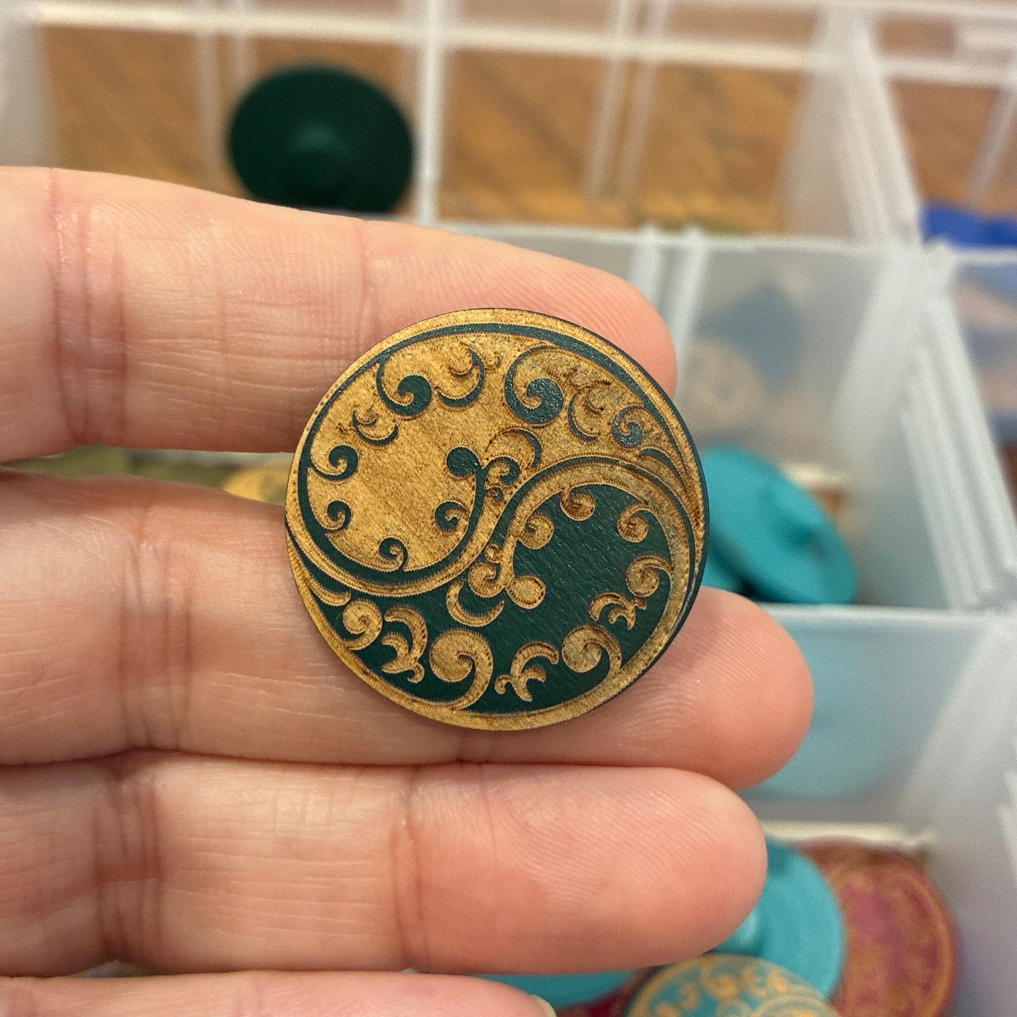 Hand holding a decorative button with intricate designs, against a background of various colored items in a storage box.