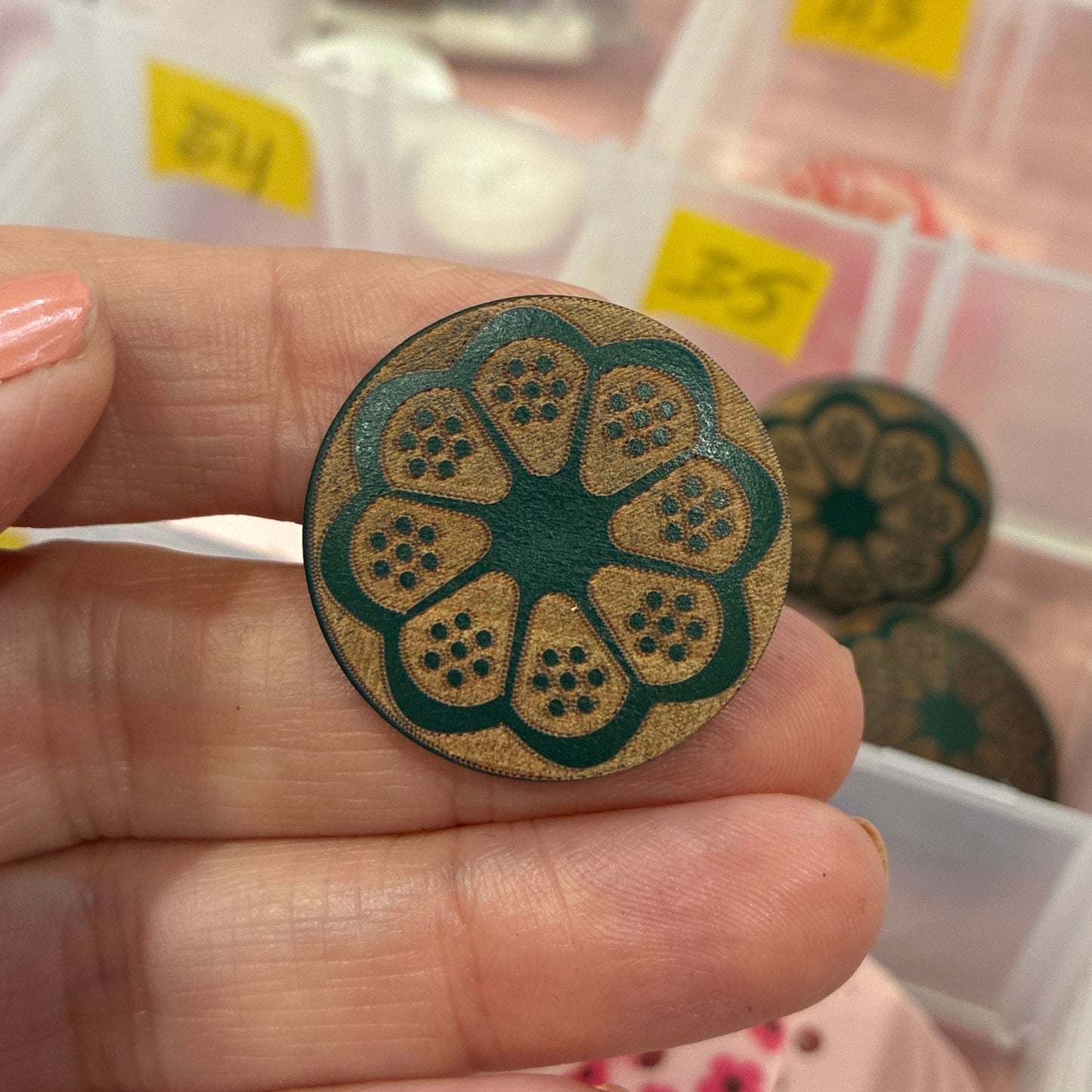 Hand holding a decorative button with a floral design in front of a container of assorted buttons.