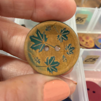 Hand holding a decorative button with leaf pattern in front of a container of buttons.