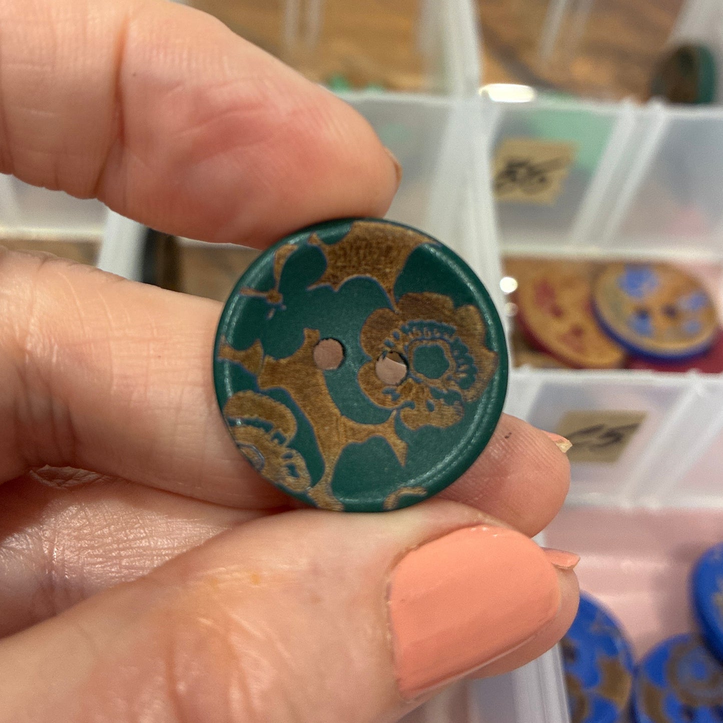 Hand holding a decorative button with intricate patterns, blurred background of a storage box with buttons.