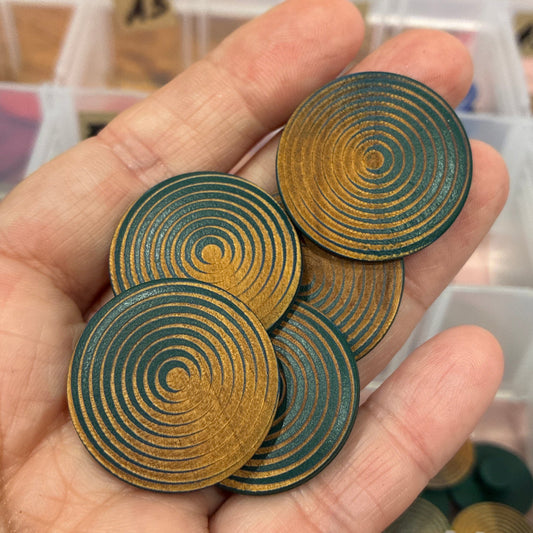 Hand holding circular wooden buttons with concentric patterns, blurred craft supplies in the background.