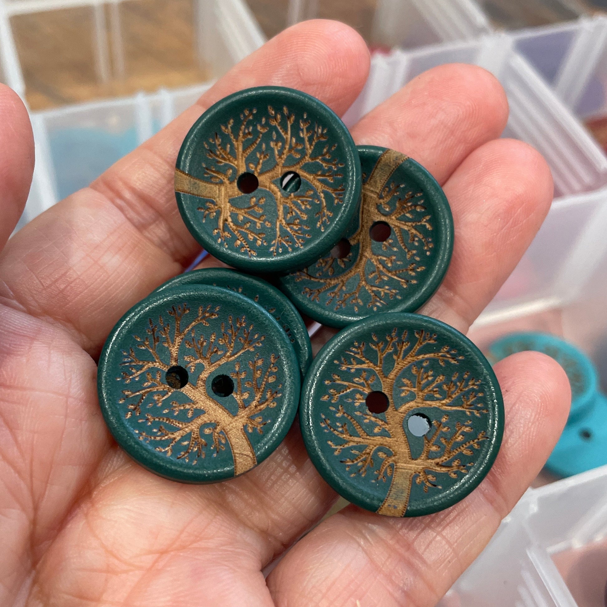 Hand holding green buttons with tree designs against a blurred background of more buttons and containers.