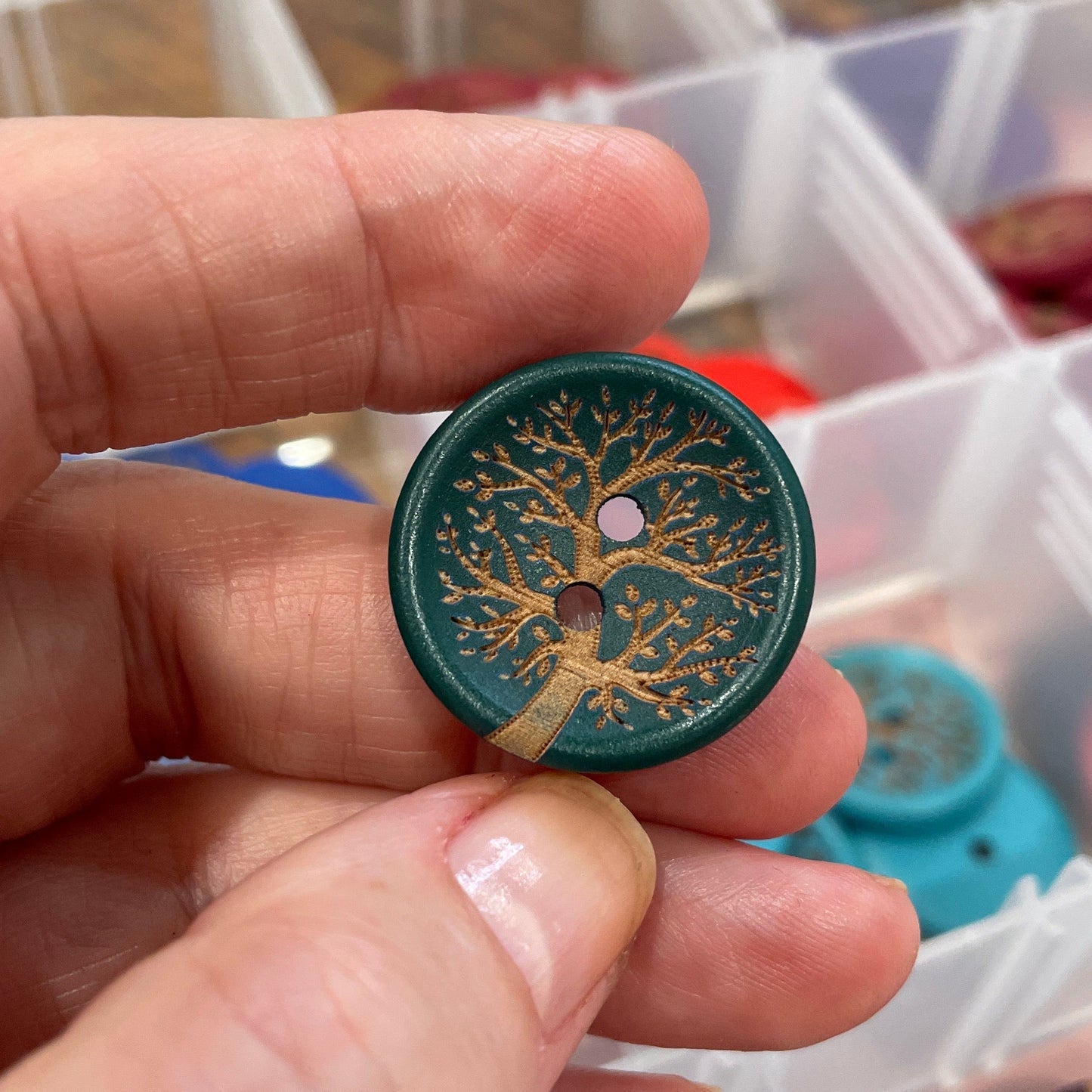 Hand holding a decorative button with a tree design, with a container of buttons in the background.