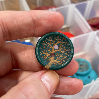Hand holding a decorative button with a tree design, with a container of buttons in the background.