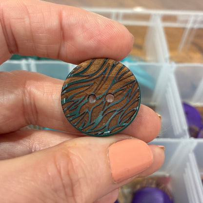 Hand holding a wooden button with a pattern, with a container of buttons in the background.
