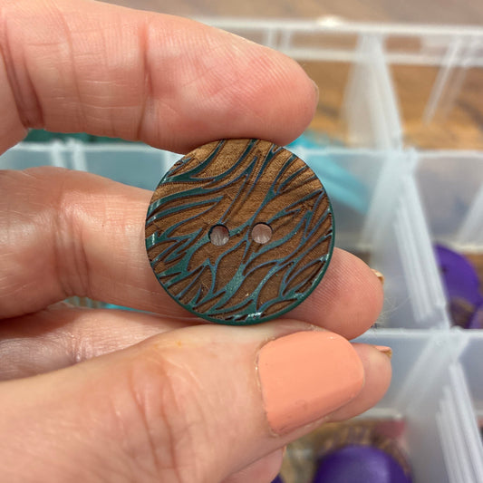 Hand holding a wooden button with a pattern, with a container of buttons in the background.