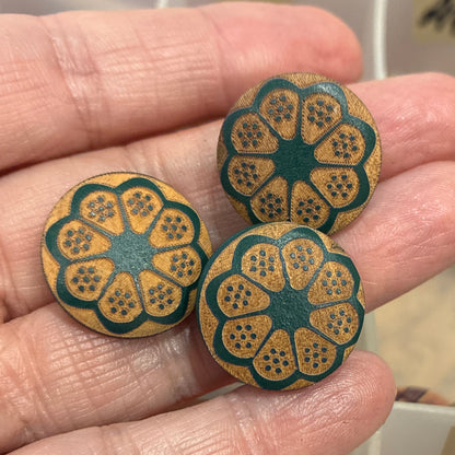 Hand holding three wooden buttons with floral design, with a container of buttons in the background.