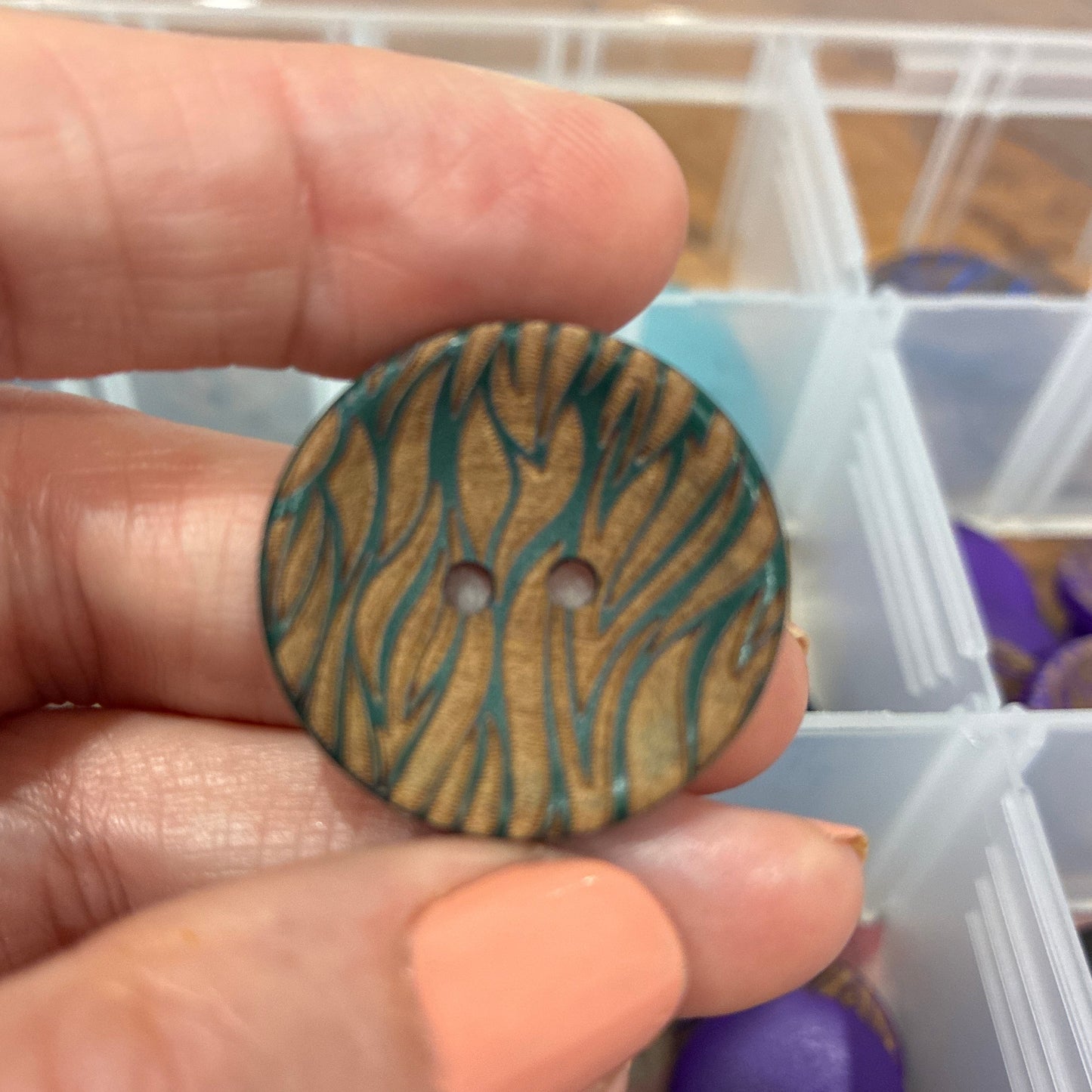 Hand holding a wooden button with a zebra pattern, with a container of buttons in the background.