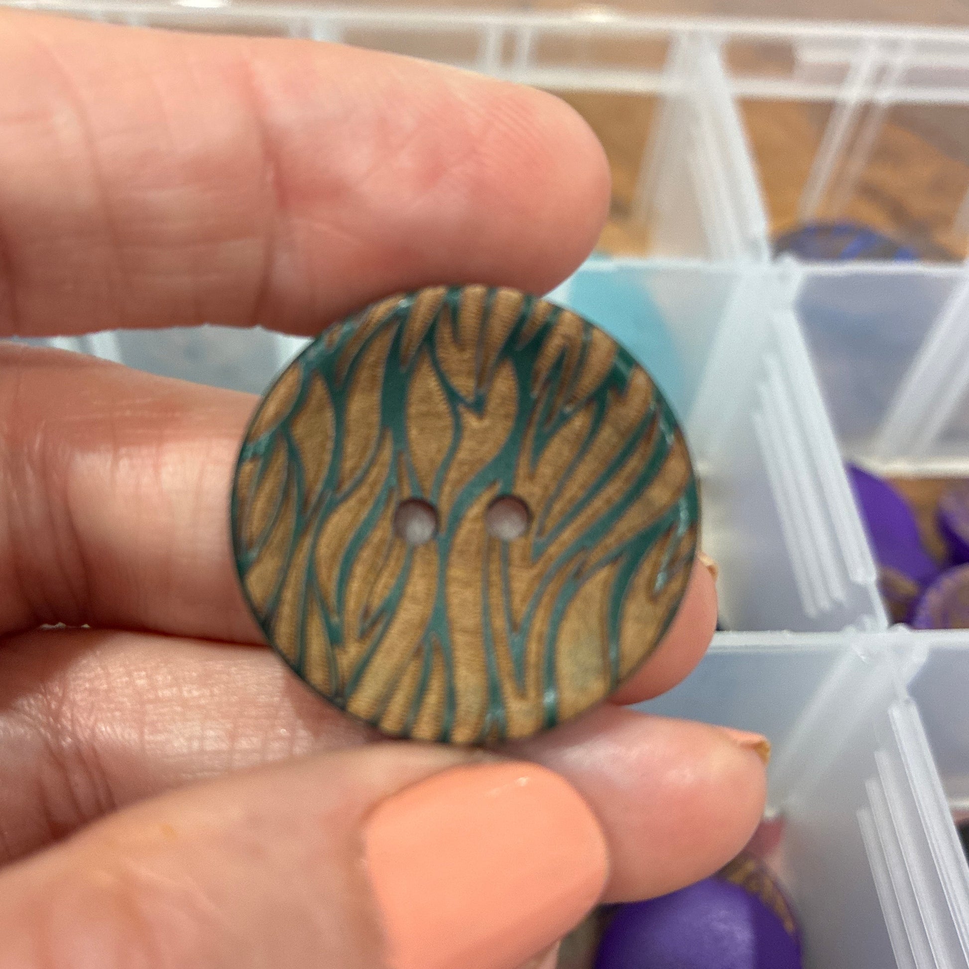 Hand holding a wooden button with a zebra pattern, with a container of buttons in the background.