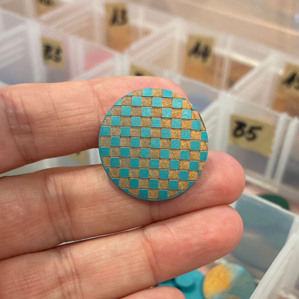 Hand holding a round, checkered button with a container of assorted items in the background
