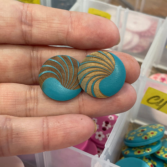 Hand holding a blue button with gold patterns in front of a container of assorted buttons.