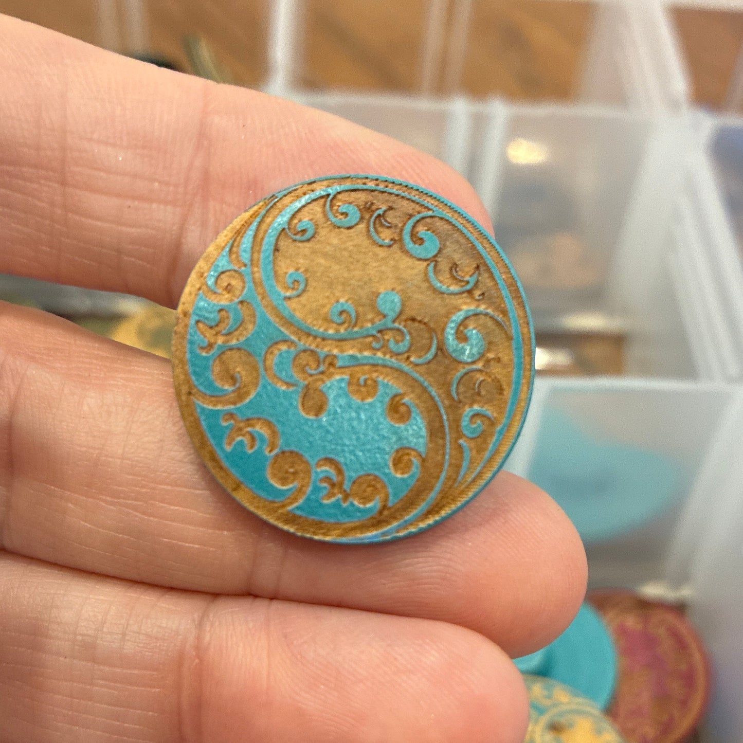 Close-up of a hand holding a decorative button with a colorful paisley design, against a blurred background of a storage box with more buttons.