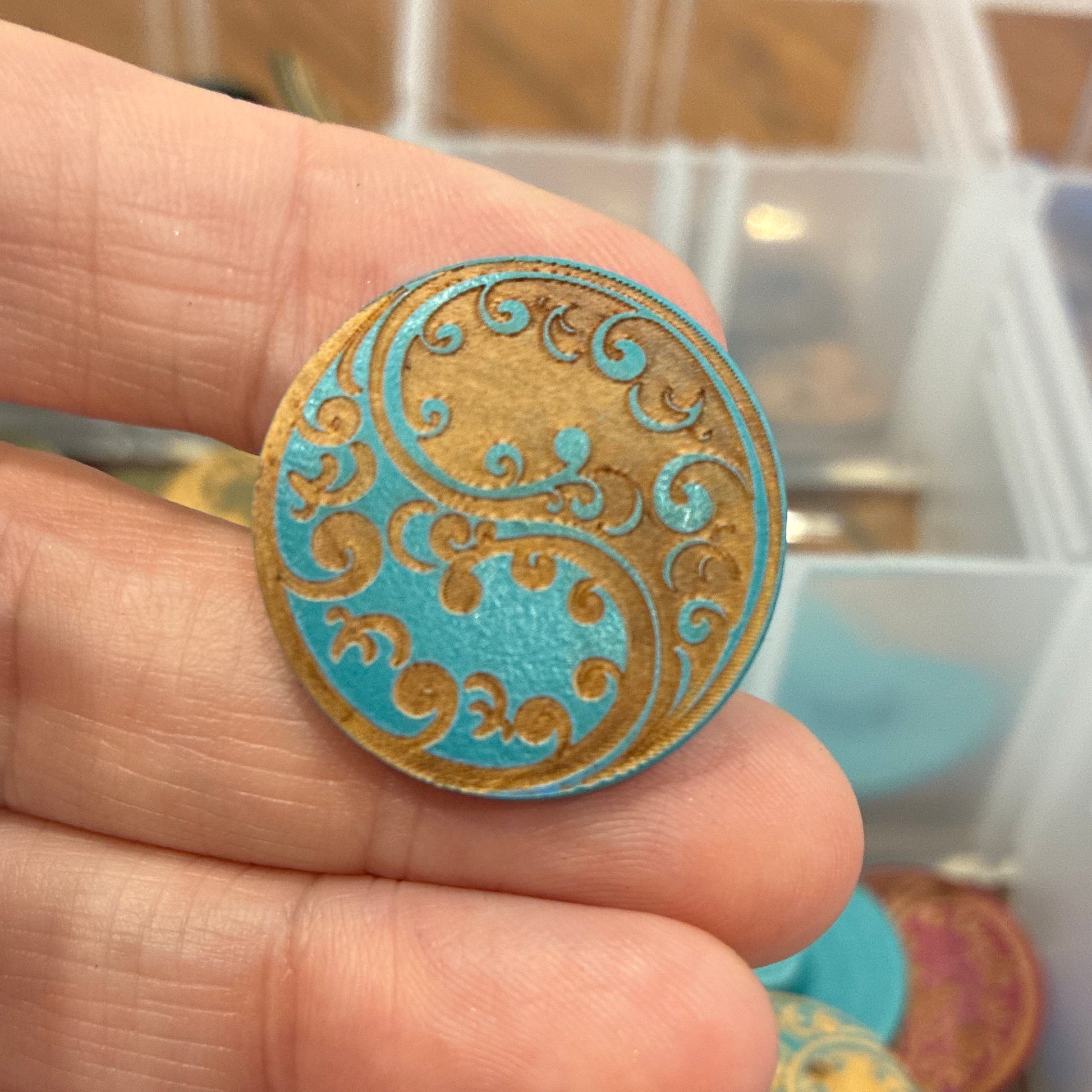 Close-up of a hand holding a decorative button with a colorful paisley design, against a blurred background of a storage box with more buttons.