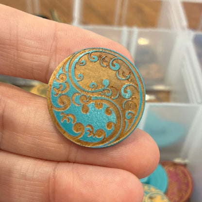 Close-up of a hand holding a decorative button with a colorful paisley design, against a blurred background of a storage box with more buttons.