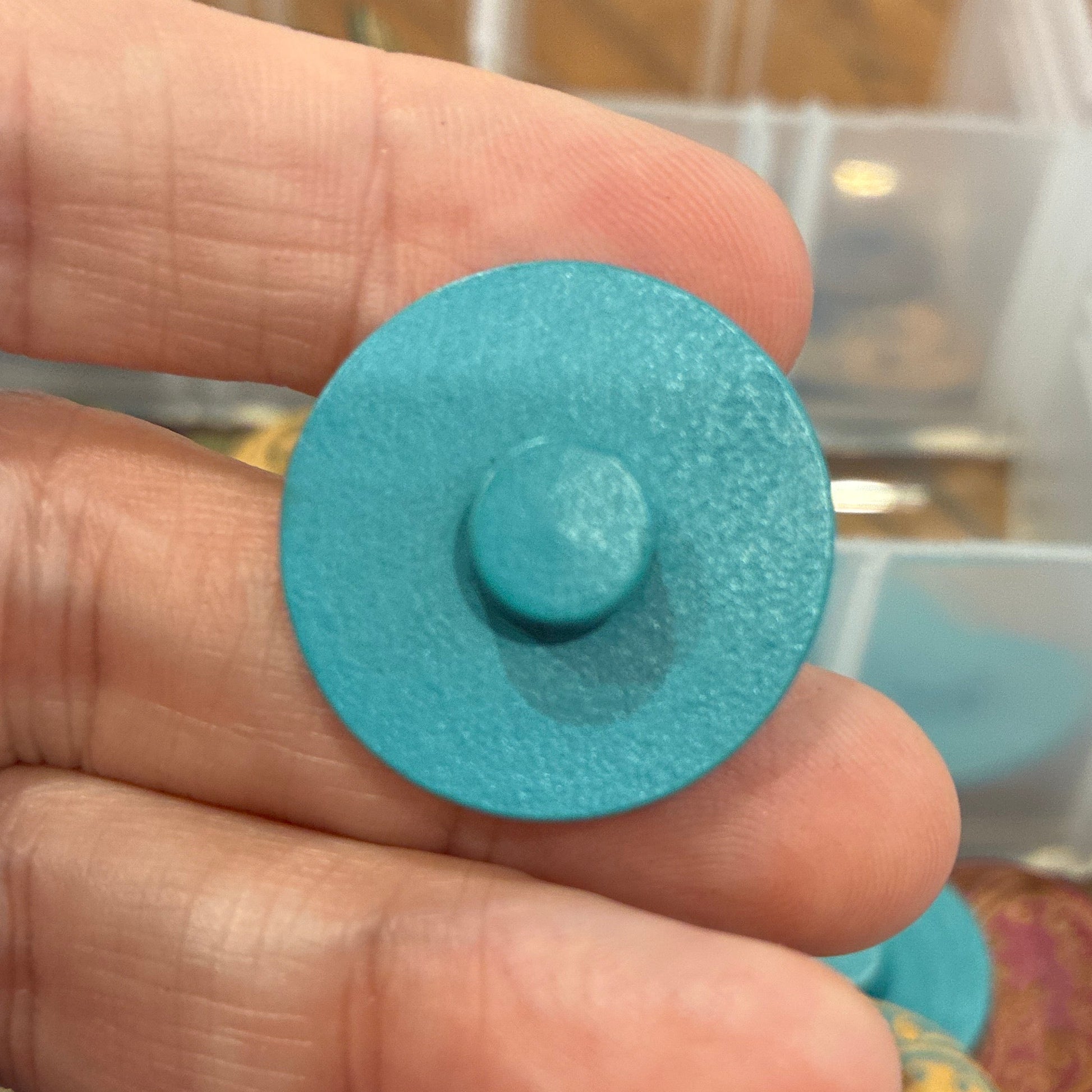 Close-up of a hand holding a blue circular object with a container of colorful objects in the background.
