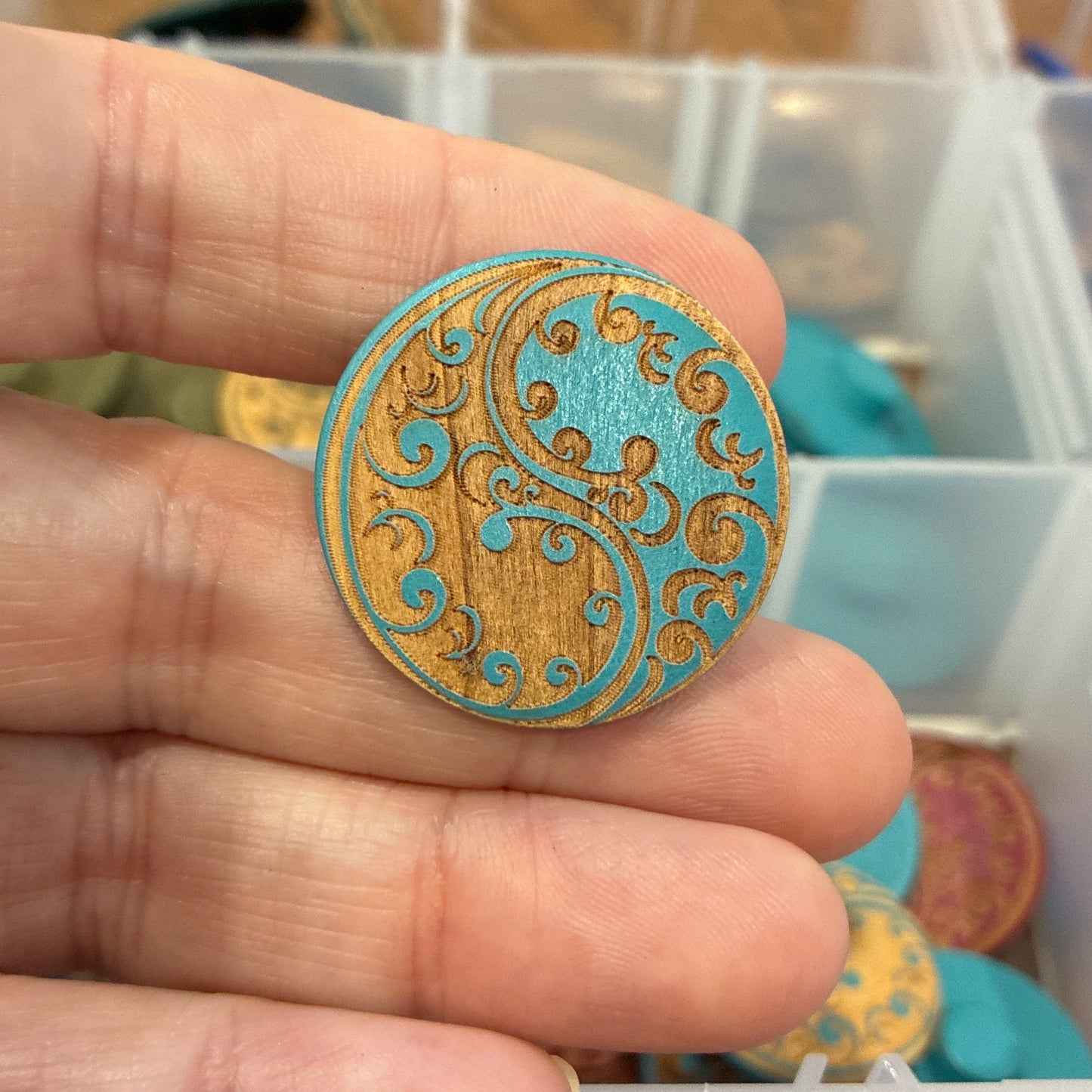 Hand holding a decorative button with intricate design, against a background of organized buttons in containers.