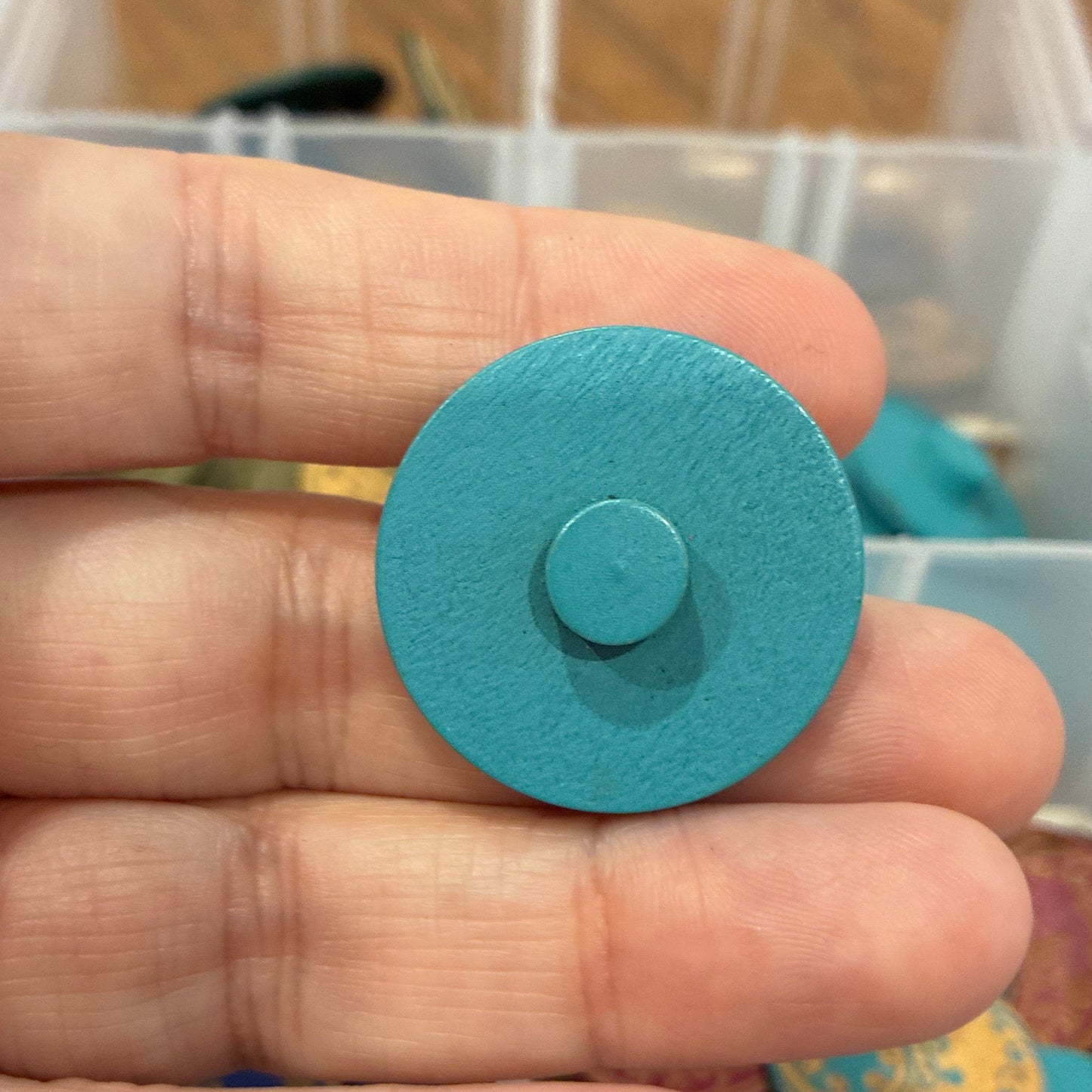 Hand holding a blue circular button with a blurred background of colorful items in a container.