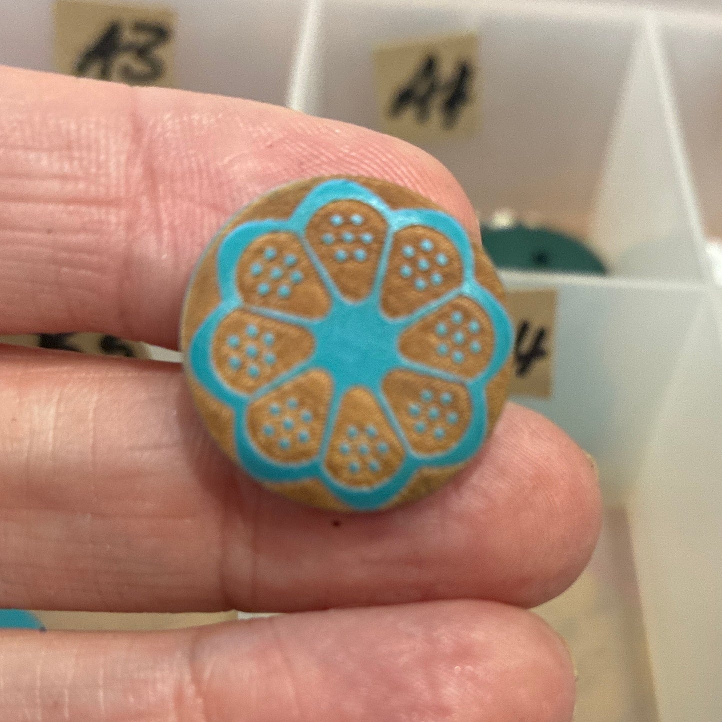 Hand holding a decorative button with a blue and gold design, with a grid of buttons in the background.