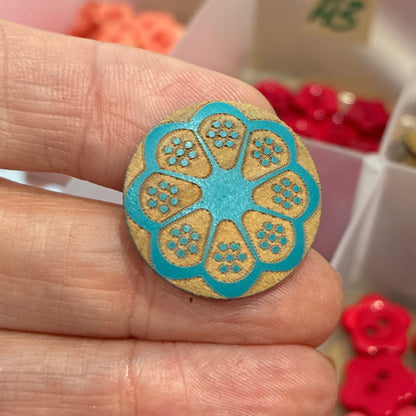 Hand holding a decorative button with a blue and gold design, with a box of buttons in the background.