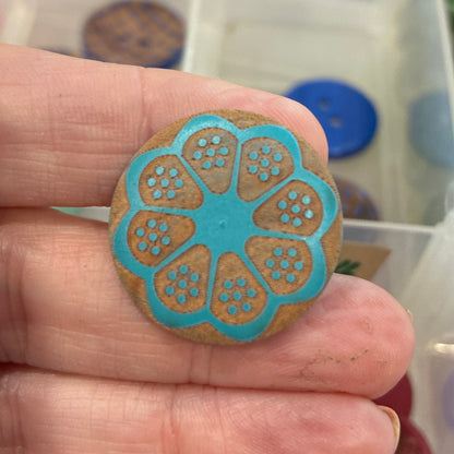 Hand holding a blue and gold decorative button with a container of buttons in the background