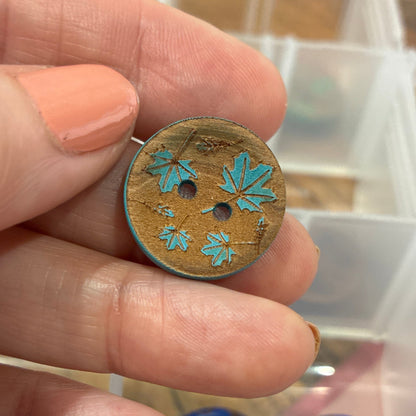 Hand holding a decorative button with floral patterns against a background of button containers.