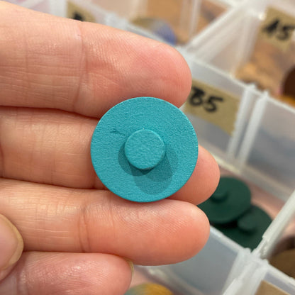 Hand holding a blue circular button with a container of assorted colored buttons  in the background.