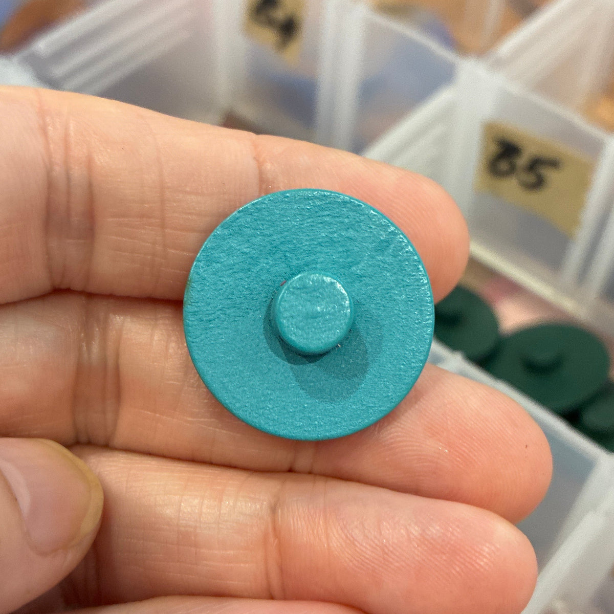 Hand holding a small blue circular button with a blurred background of a storage box with compartments.