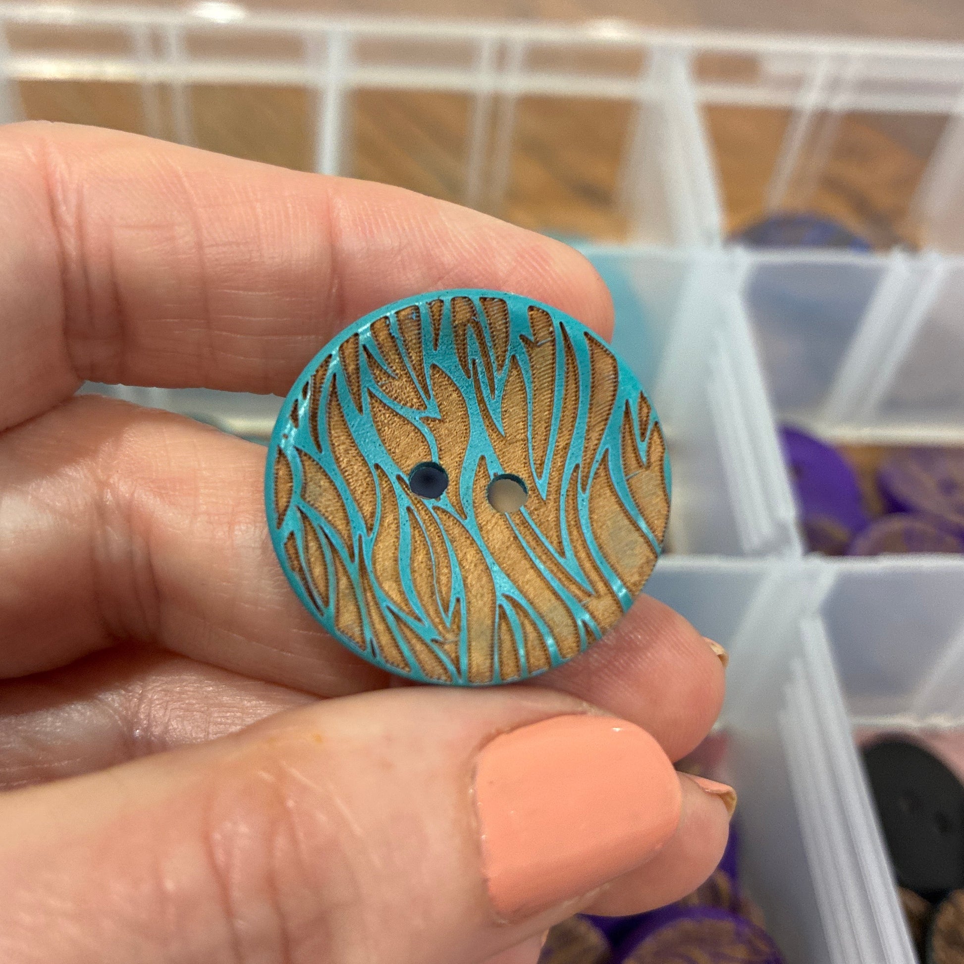 Hand holding a blue and brown patterned button with a clear container of buttons in the background.