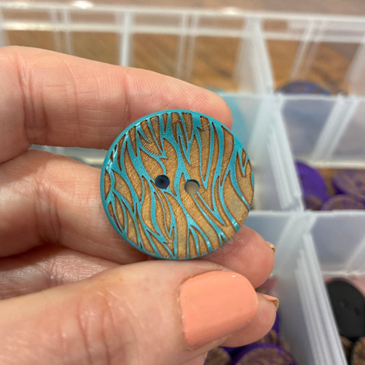 Hand holding a blue and brown patterned button with a clear container of buttons in the background.