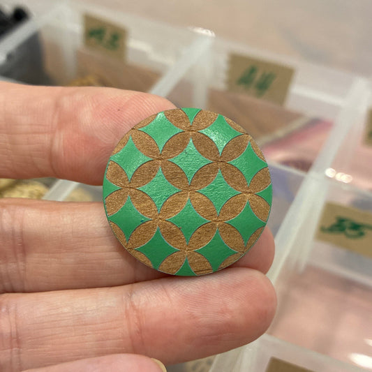 Hand holding a green and gold decorative button with a container of buttons in the background.