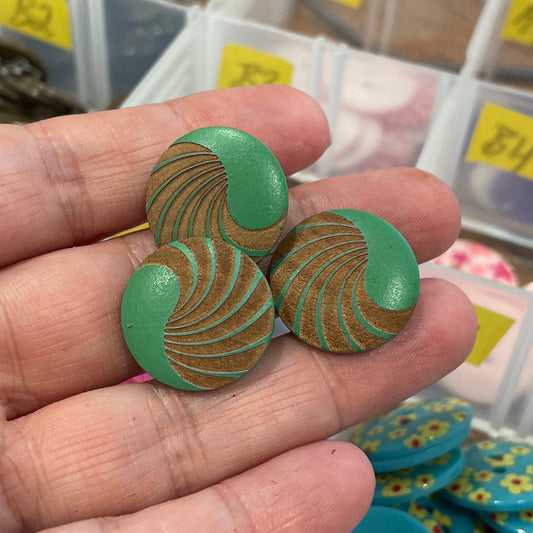 Hand holding green buttons with a background of button storage containers.