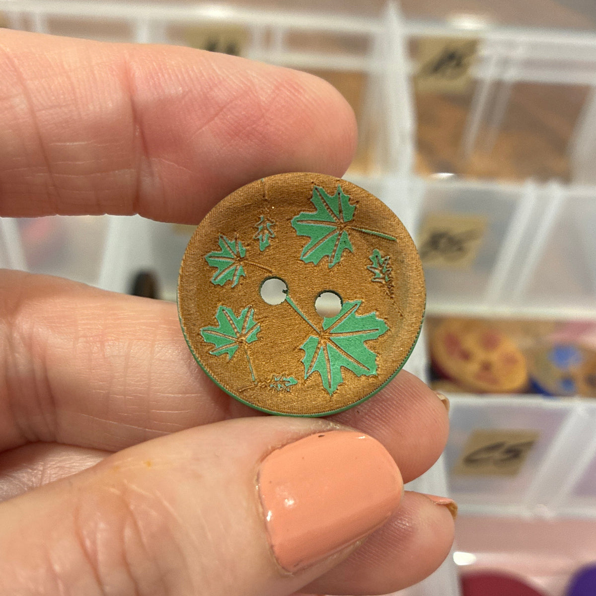 Hand holding a decorative button with green leaf patterns in front of a container of buttons.