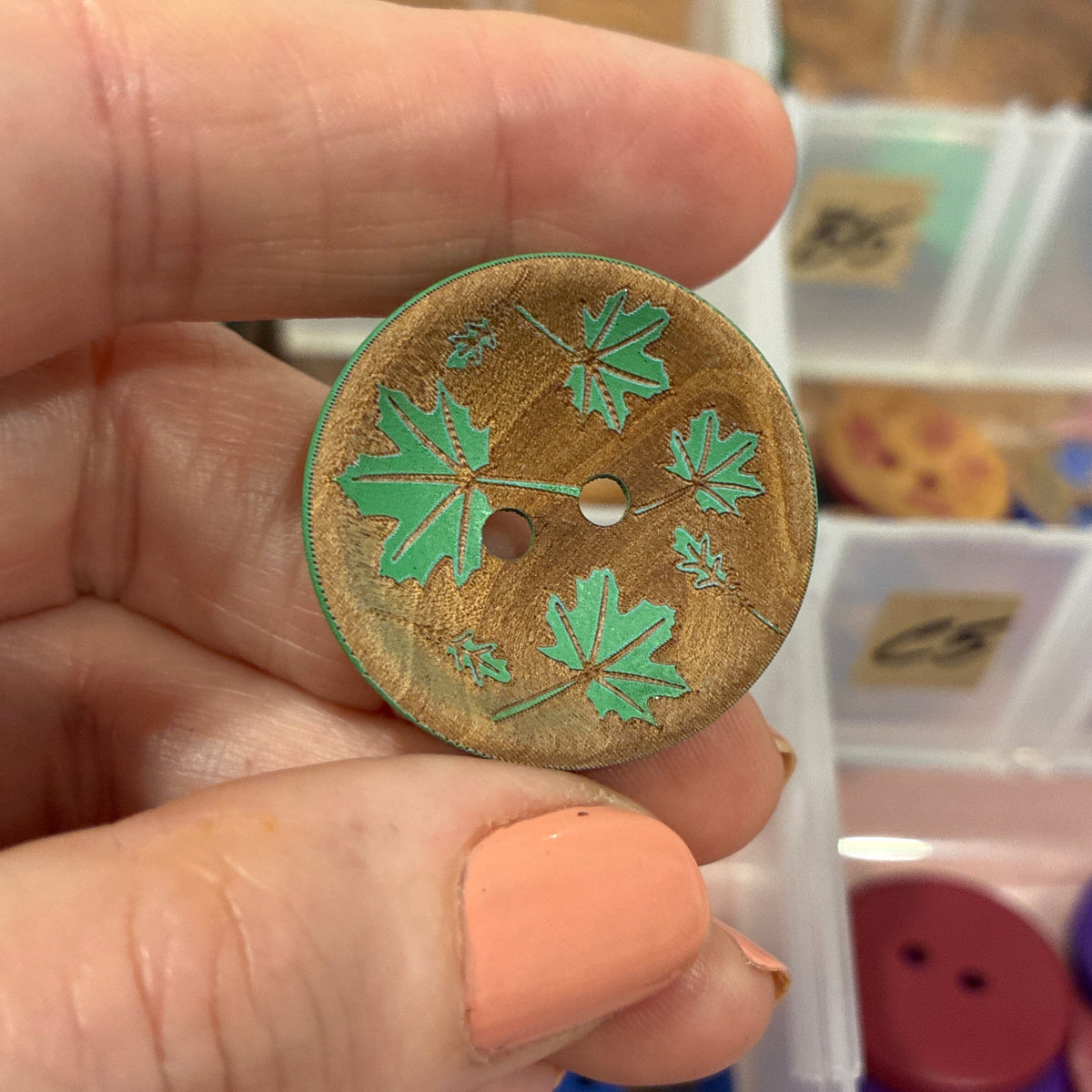 Hand holding a decorative button with green leaf design in front of a container of buttons.