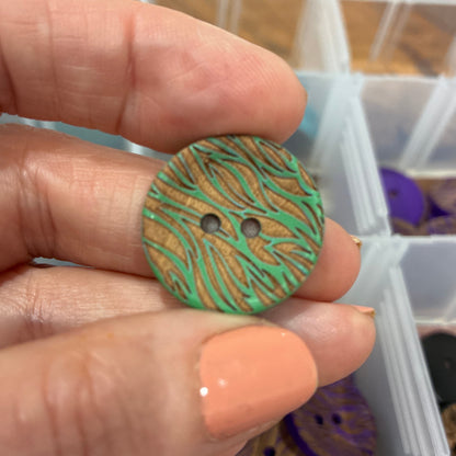 Hand holding a green patterned button with a container of buttons in the background