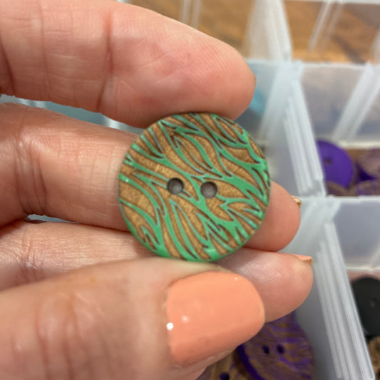Hand holding a green patterned button with a container of buttons in the background