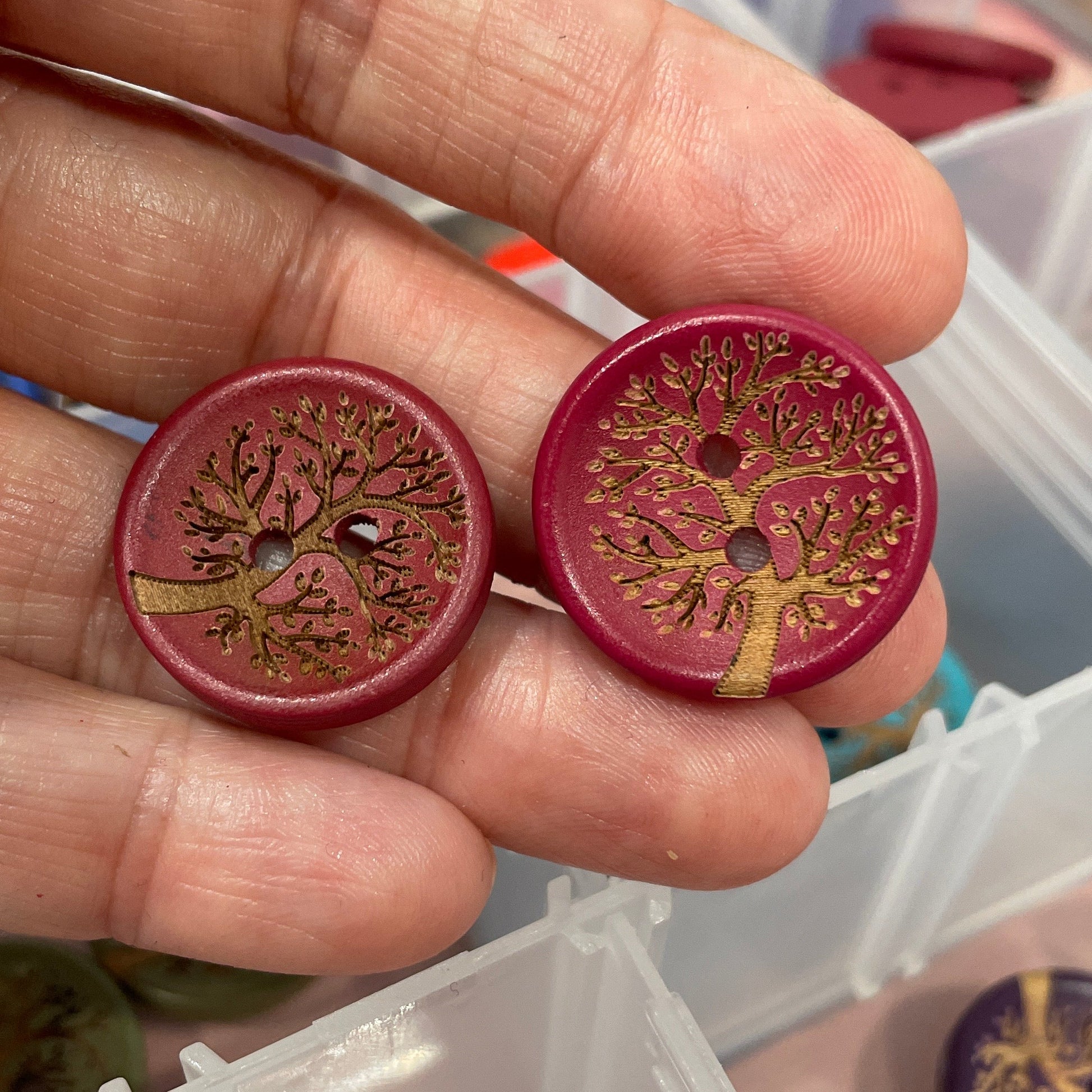 Hand holding two decorative buttons with intricate designs, with a blurred background of containers.