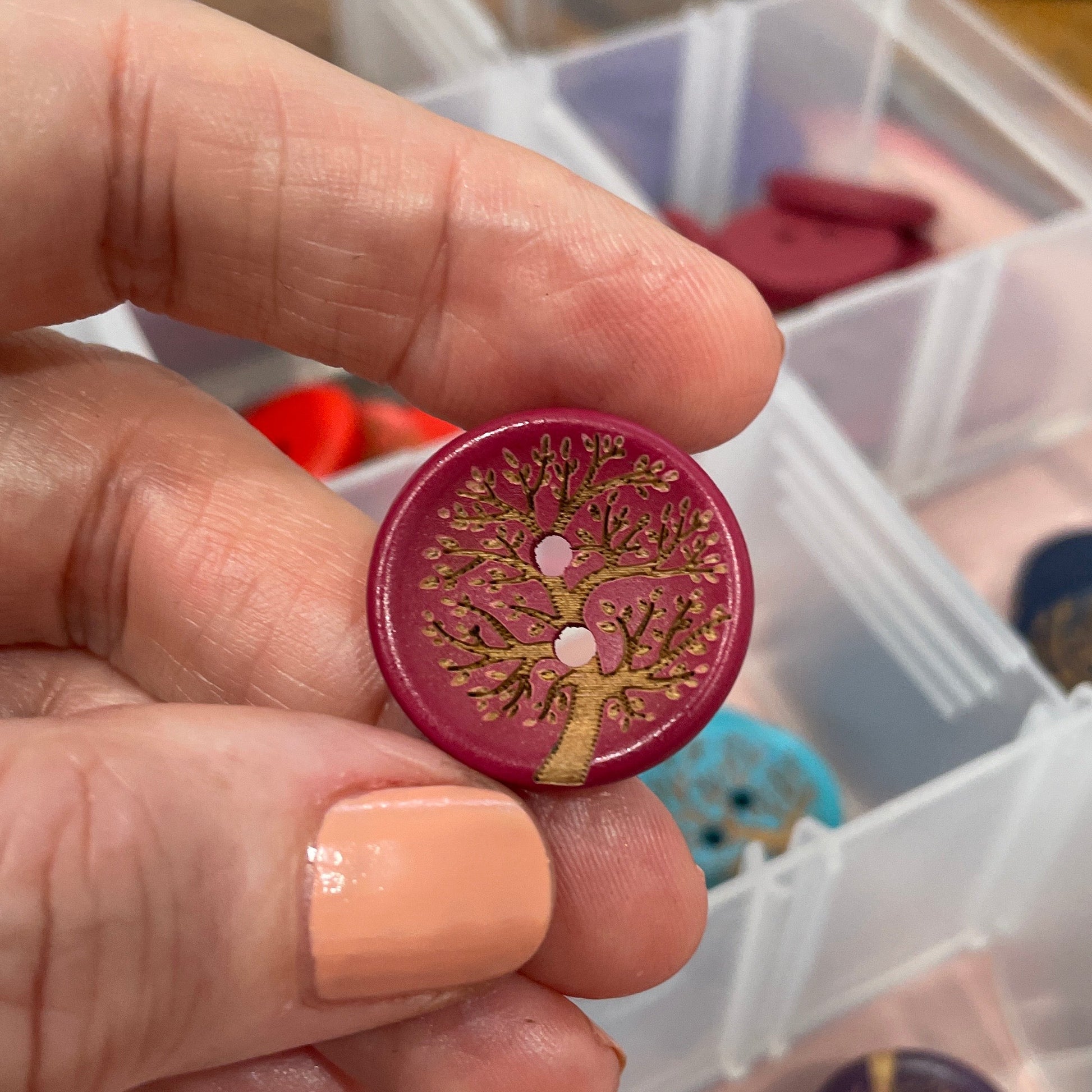 Hand holding a decorative button with intricate design, surrounded by a clear container with more buttons.