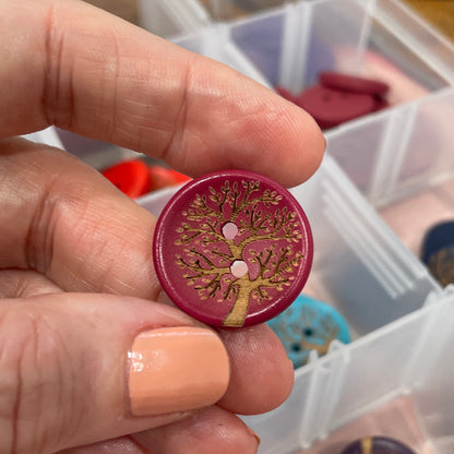 Hand holding a decorative button with intricate design, surrounded by a clear container with more buttons.